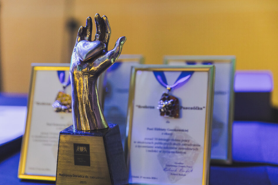 na zdjęciu metalowa statuetka w kształcie ludzkiej ręki, i kilka oszklonych papierowych dyplomów, stoją na stoliku