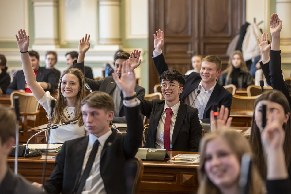 na zdjęciu kilkunastu młodych ludzi, siedzą w kilku rzędach za brązowymi stołami, podnoszą ręce w górę w trakcie prawdopodobnie głosowania