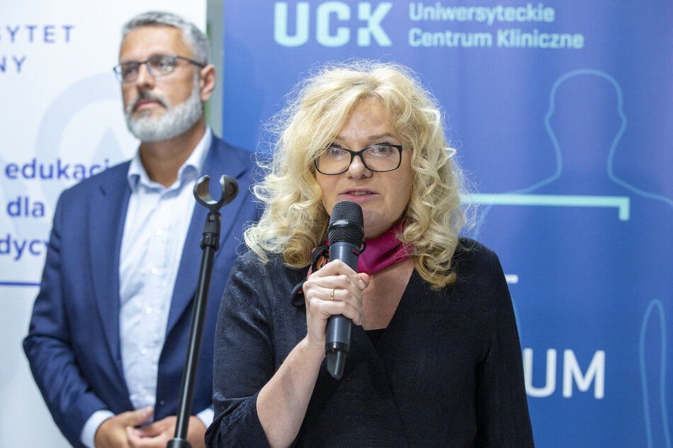 kobieta mówiąca do mikrofonu na konferencji. Ma na sobie czarną bluzę i różowy szalik na szyi. Za nią jest mężczyzna z białą brodą i okularami