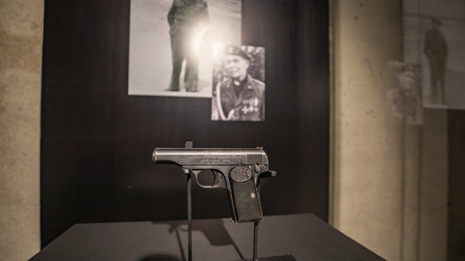 pistolet z czasów II wojny światowej na wystawie w ciemnym otoczeniu, w tle zdjęcia mjr. Sucharskiego