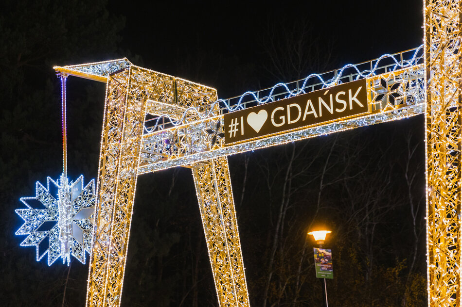 duża metalowa konstrukcja z napisem GDANSK w białych literach. Konstrukcja jest ozdobiona niebieskimi i białymi światłami, tworząc świąteczną i radosną atmosferę. Po lewej stronie wisi niebieska dekoracja w kształcie gwiazdy