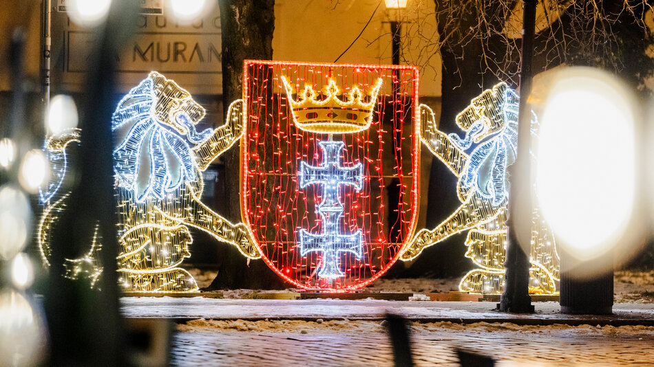  Duża czerwona tarcza ze złotą koroną na górze, która jest oświetlona niebieskimi i białymi światłami. Tarcza jest w środku otoczona dwoma lwami po obu stronach. Nad tarczą znajduje się niebieski krzyż i baner z literami H i C na niej. W tle możemy zobaczyć ulicę z drzewami oraz budynkami, a na chodniku jest kilka osób.