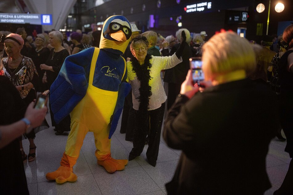 Seniorka pozuje do zdjęcia z niebiesko-żółtą maskotką gdańskiego lotniska. W tle tańczący i bawiący się ludzie