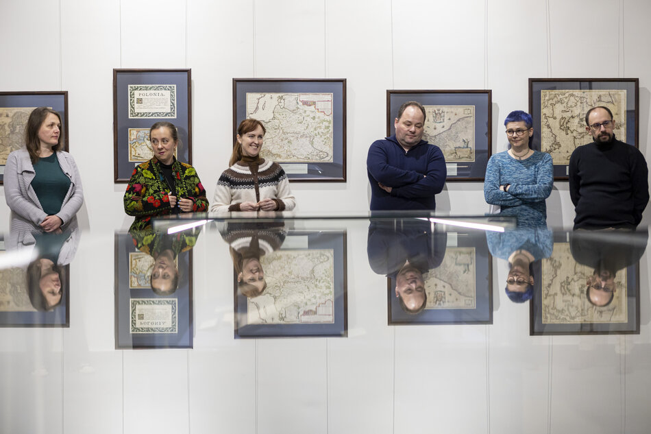 Sześć osób uczestniczy w wernisażu wystawy map, prawdopodobnie związanej z historią Polski lub kartografią. Ekspozycja odbywa się w historycznym gmachu PAN Biblioteki Gdańskiej i prezentuje nowe nabytki jej zbiorów specjalnych, w tym wiekową mapę Pomorza z 1573 roku.