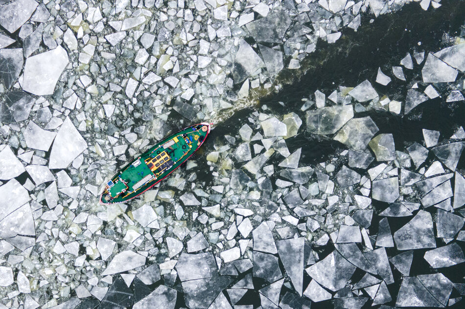 Zdjęcie z lotu ptaka. Lodołamacz płynie przez zamarzniętą rzekę, zostawiając za sobą udrożniony tor.