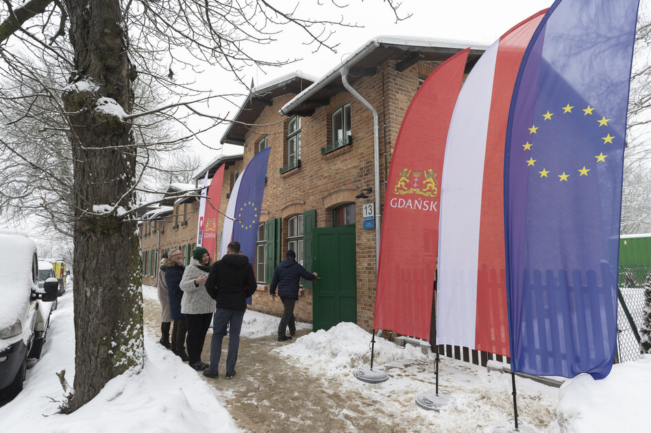 Ceglany dom w śnieżnej scenerii. Przed domem flagi Gdańska, Polski i Unii Europejskiej oraz kilka osób.