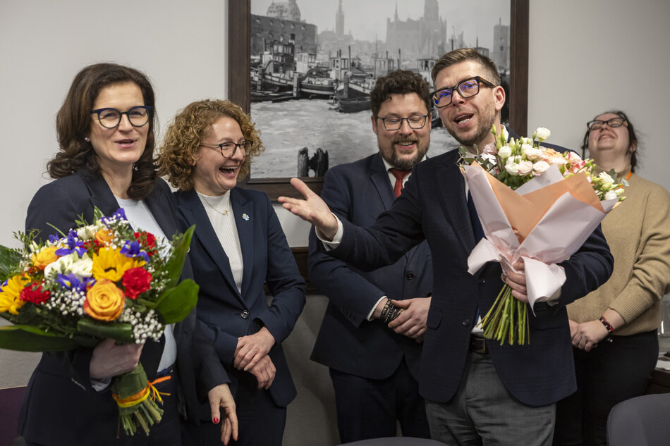 Mężczyzna trzyma kwiaty i się śmieje, obok dwie kobiety się śmieją, jedna z nich trzyma bukiet kwiatów.