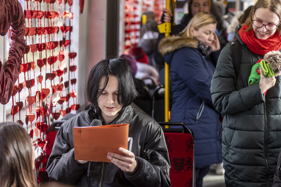 W centralnym punkcie zdjęcia znajduje się młoda osoba o ciemnych włosach w skórzanej kurtce, która z uwagą czyta tekst z pomarańczowej teczki. W tle widoczni są inni pasażerowie tramwaju, a wnętrze pojazdu zdobią liczne czerwone serca, tworząc odświętną, walentynkową atmosferę.