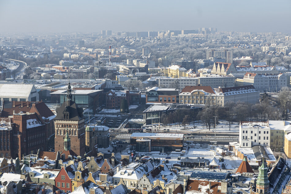 Na zdjęciu widać zimową panoramę Gdańska, wykonaną prawdopodobnie z wieży Bazyliki Mariackiej, ukazującą dachy kamienic pokryte śniegiem. W centrum uwagi znajduje się zabytkowa Wieża Więzienna oraz Katownia, które stanowią część historycznego Zespołu Przedbramia przy ulicy Długiej