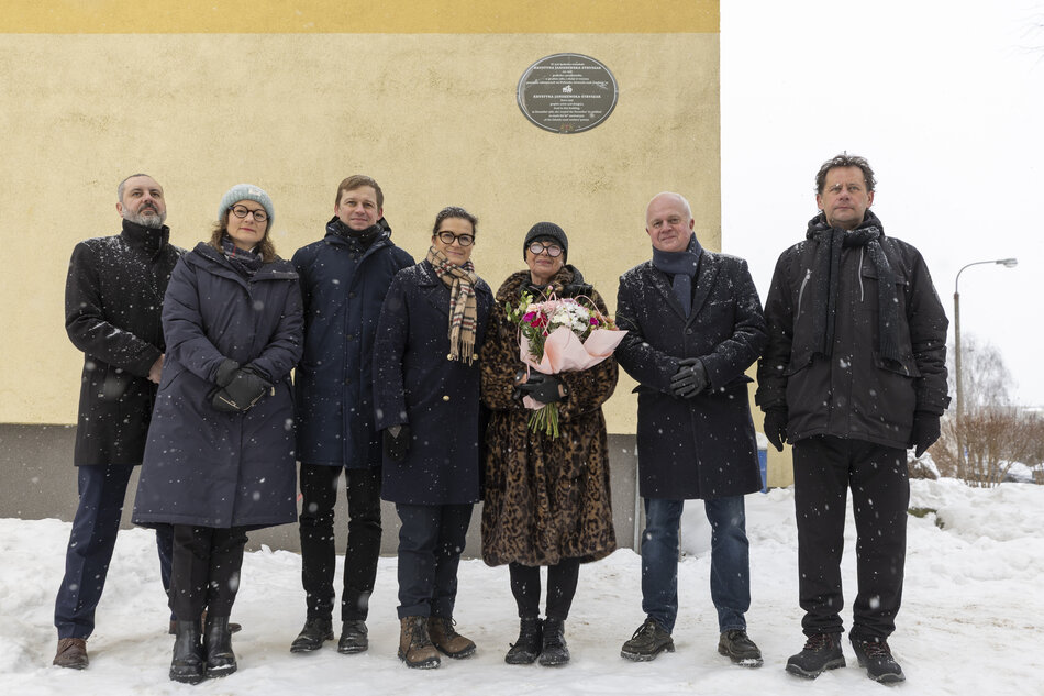 Na zdjęciu widać grupę osób uczestniczących w uroczystym odsłonięciu tablicy pamiątkowej na ścianie budynku. Zgromadzeni, ubrani w zimowe stroje, trzymają czerwoną wstęgę i bukiet kwiatów, a cała scena rozgrywa się w zimowej scenerii na tle ośnieżonego terenu.