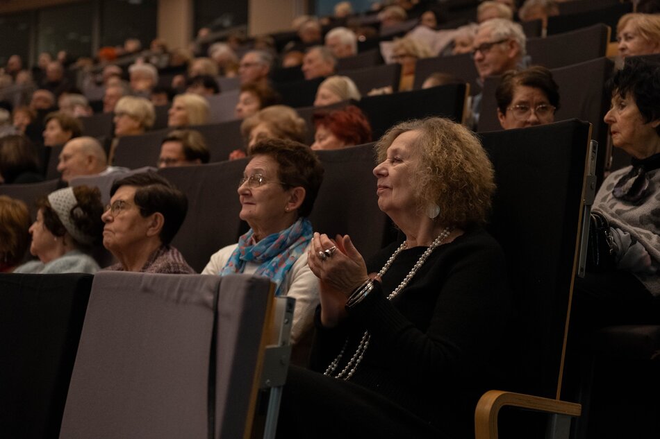 Zdjęcie przedstawia liczną widownię, składającą się głównie z osób starszych, siedzącą w ciemnej sali wykładowej lub teatralnej. Uczestnicy z uwagą obserwują wydarzenie, a kobieta na pierwszym planie uśmiecha się i klaszcze, co sugeruje pozytywny odbiór prezentacji lub występu. 