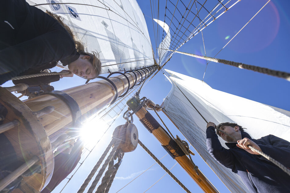 Zdjęcie przedstawia załogę pracującą przy olinowaniu i żaglach tradycyjnego żaglowca na tle bezchmurnego nieba. Dynamiczne ujęcie z dołu eksponuje drewniane maszty, bloki oraz napięte płótna w pełnym słońcu, oddając atmosferę aktywnej żeglugi.