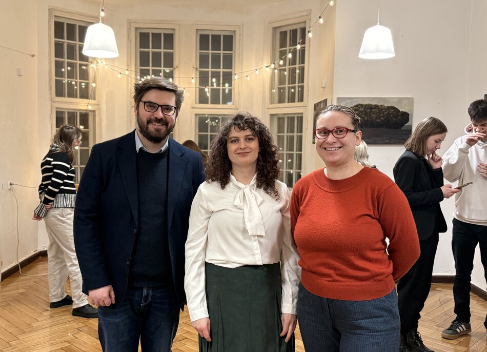 Zdjęcie przedstawia grupę osób uśmiechających się do aparatu w jasnym pomieszczeniu ozdobionym sznurem lampek. Na pierwszym planie widać mężczyznę w ciemnej marynarce oraz dwie kobiety – jedną w białej bluzce z wiązaniem, a drugą w czerwonym swetrze. 