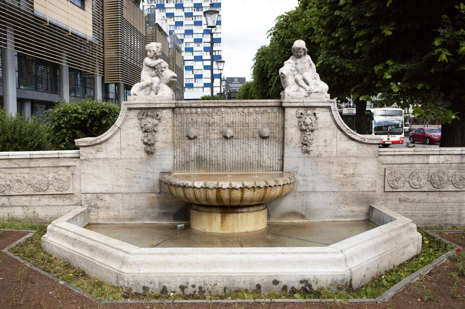 na zdjęciu jasna niewysoka fontanna, w tle po lewej i z tyłu budynki