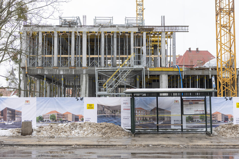 na zdjęciu budowany budynek o betonowej konstrukcji, przed nim ogrodzenie i wiata przystankowa