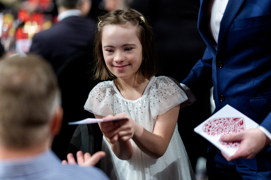 Na zdjęciu widać uśmiechniętą dziewczynkę w błyszczącej, białej sukience, która wręcza komuś kartę lub małą kopertę. Obok niej stoi mężczyzna w niebieskiej marynarce, trzymający w dłoniach przedmiot przypominający talię kart lub ozdobne zaproszenie. 