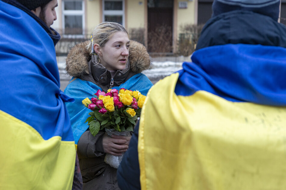 Trzy osoby otulone flagami Ukrainy, kobieta trzyma bukiet kwiatów.
