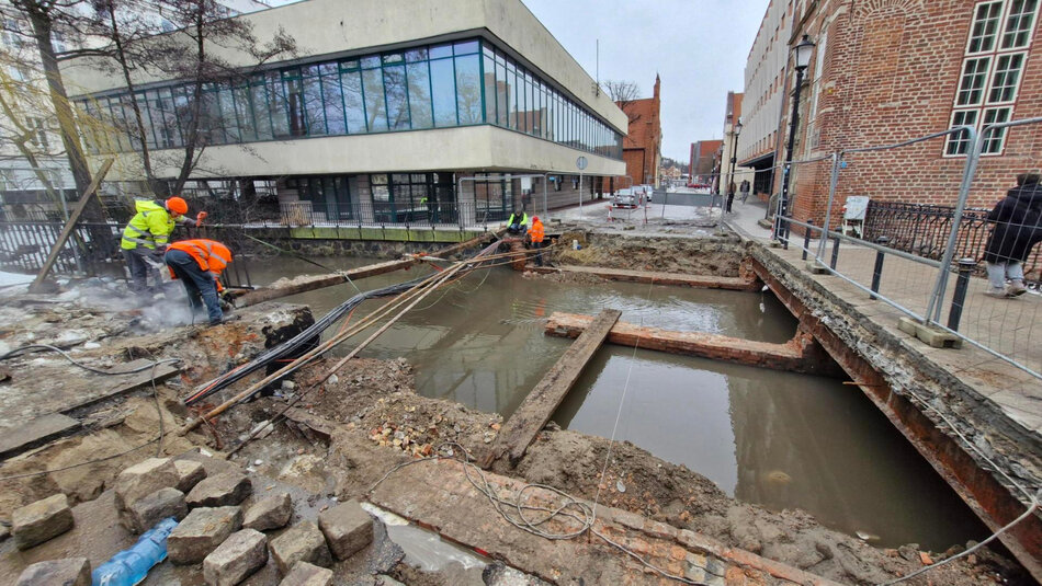 na zdjęciu miejsce po rozebranym moście, widać plac budowy, brzegi rzeki, w tle budynki po obu stronach ulicy