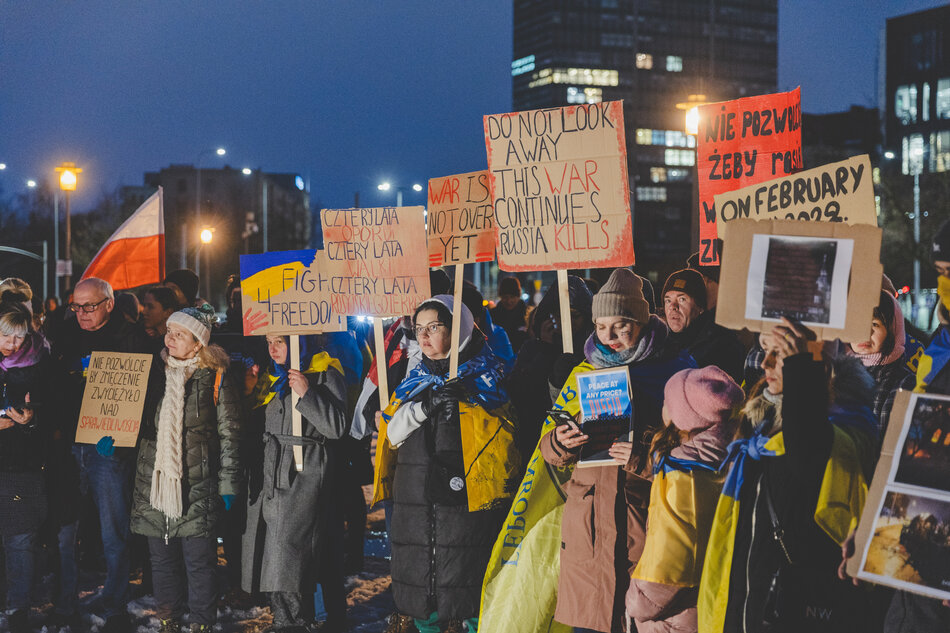 Na zdjęciu widać grupę ludzi biorących udział w proteście lub manifestacji, trzymających flagi Ukrainy oraz transparenty z napisami w języku angielskim.