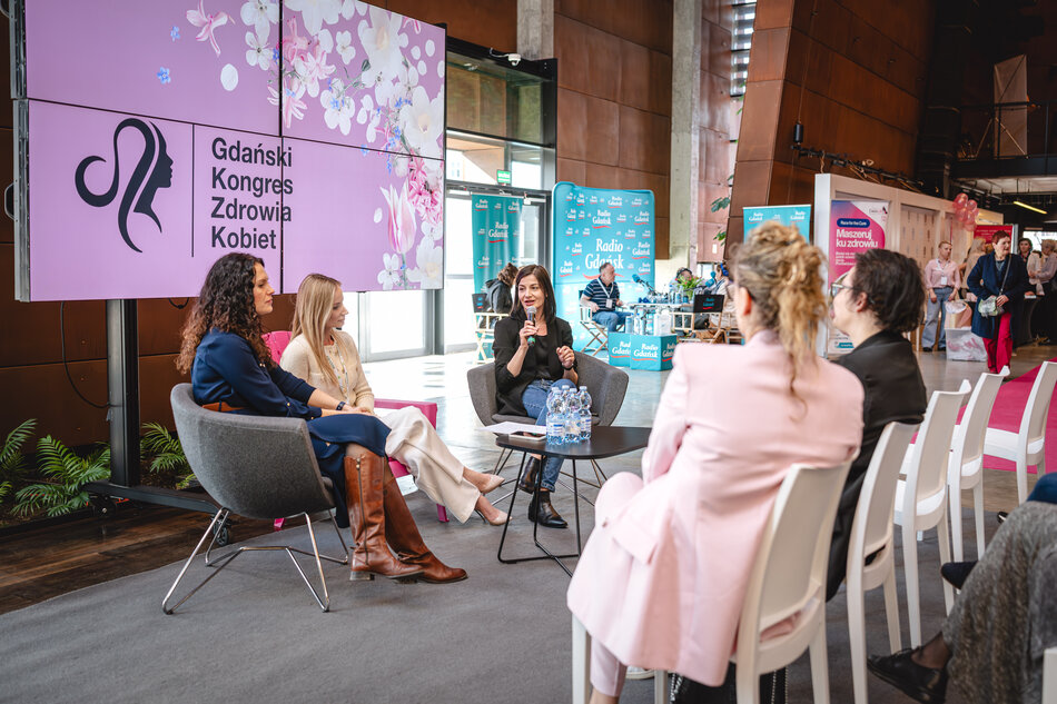 Trzy kobiety siedzą w fotelach na scenie podczas panelu dyskusyjnego na wydarzeniu „Gdański Kongres Zdrowia Kobiet”. Jedna z nich trzyma mikrofon i przemawia, pozostałe słuchają. W tle widać stoisko Radia Gdańsk oraz uczestników konferencji, a na dużym ekranie obok sceny widnieje logo i nazwa wydarzenia na różowym tle z motywem kwiatowym.