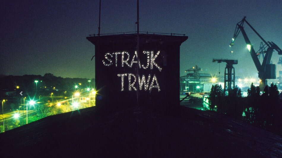Nocne zdjęcie terenu stoczni zrobione z poziomu dachów hal produkcyjnych. Na pierwszym planie na dachu wielki napis białą farbą Strajk Trwa. W tle panorama miasta i dźwigi stoczniowe