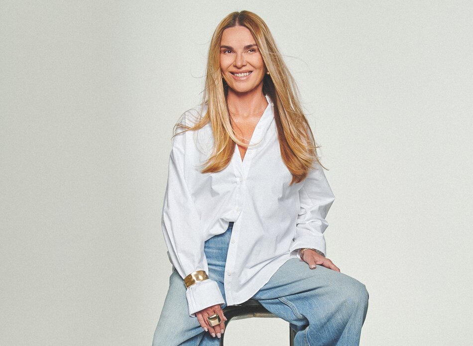 Na zdjęciu widać uśmiechniętą kobietę, dziennikarkę Hannę Lis, siedzącą na wysokim metalowym stołku na jasnym, jednolitym tle. Ma długie, proste blond włosy i naturalny makijaż. Ubrana jest w luźną, białą koszulę z długim rękawem oraz jasnoniebieskie, szerokie dżinsy z wysokim stanem. Na nadgarstku ma szeroką, złotą bransoletę i pierścionek. Całość ma minimalistyczny, studyjny charakter.