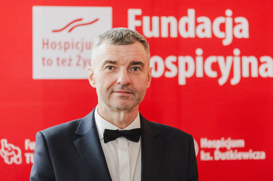 Na portretowym zdjęciu widoczny jest mężczyzna w ciemnym garniturze i eleganckiej muszce, który spogląda bezpośrednio w obiektyw. Tło stanowi czerwona ściana z białymi logotypami Fundacji Hospicyjnej oraz Hospicjum ks. Dutkiewicza.