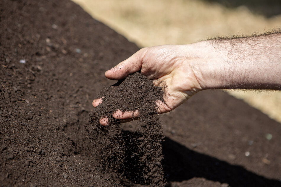 na pierwszym planie ręka. Z jej dłoni wysypuje się ciemna ziemia. W tle znajduje się góra ziemi dominująca kadr