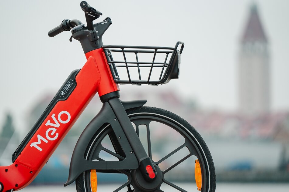 Na zdjęciu widać czerwony rower elektryczny marki Segway, będący częścią systemu roweru metropolitalnego Mevo w Obszarze Metropolitalnym Gdańsk-Gdynia-Sopot. Pojazd jest wyposażony w czarny przedni koszyk, charakterystyczne pełne obręcze kół oraz opony z elementami odblaskowymi