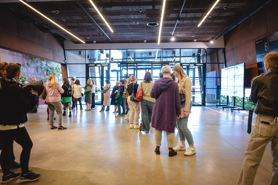 Grupa osób stoi i rozmawia w holu Europejskiego Centrum Solidarności. Wnętrze ma betonową podłogę, ciemny sufit z podłużnymi lampami LED oraz rośliny przy ścianach. Po prawej stronie widoczny jest duży ekran informacyjny.