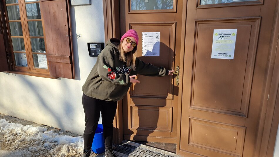 Dziewczyna w różowej wełnianej czapce i okularach otwiera drzwi w zapraszającym geście