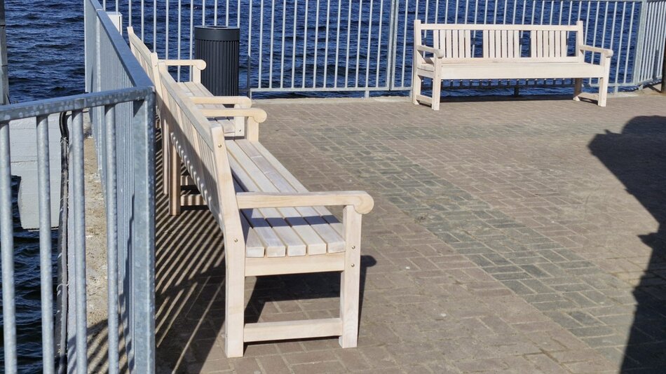 Na pierwszym planie, obok białych barierek, znajduje się biała ławka. W tle widać kolejną, a za nią niebieskie morze. Ławki stoją na podeście zrobionym z kostki brukowej