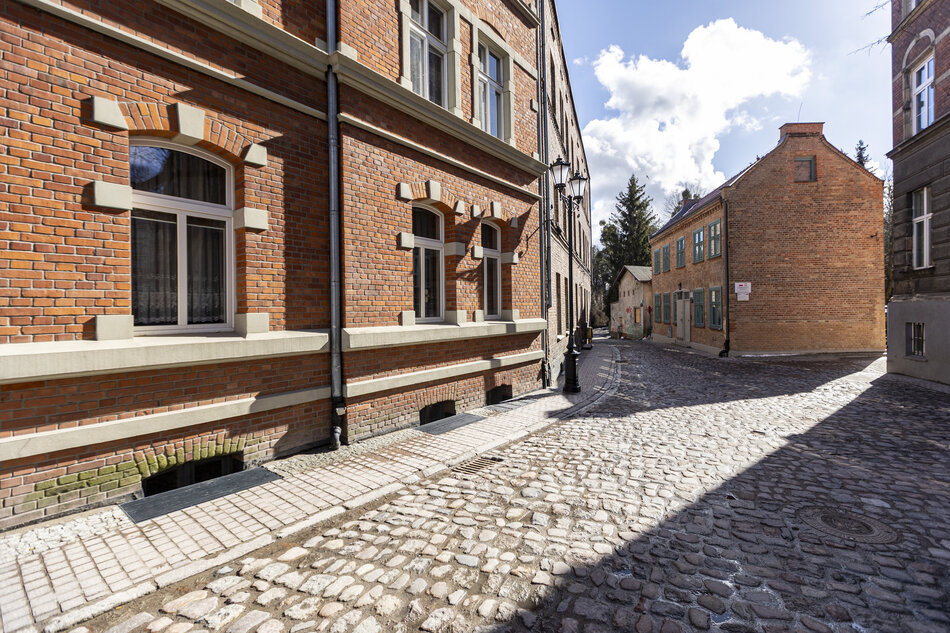 Zdjęcie przedstawia urokliwą, brukowaną uliczkę biegnącą wzdłuż zabytkowych kamienic wykonanych z czerwonej cegły. Charakterystyczna architektura i detale, takie jak stylowe latarnie oraz okna z jasnymi obramowaniami, tworzą klimat dawnego Gdańska na Biskupiej Górce.