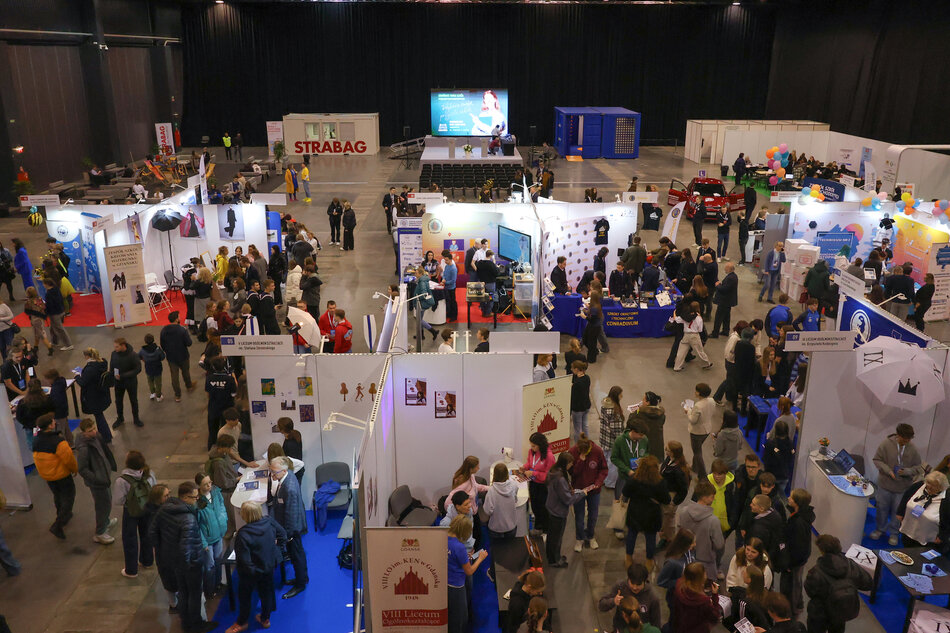 duża sala wystawowa wypełniona ludźmi. Sala jest ozdobiona białymi i niebieskimi banerami i stoiskami, a w całej przestrzeni rozrzuconych jest kilka stołów i foteli.