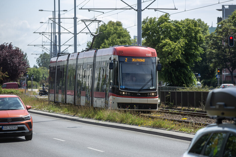 czerwono-biały tramwaj, porusza się wzdłuż torów i wydaje się być w ruchu. Ma numer 2 z przodu. Obok niego, po jezdni, jadą samochody