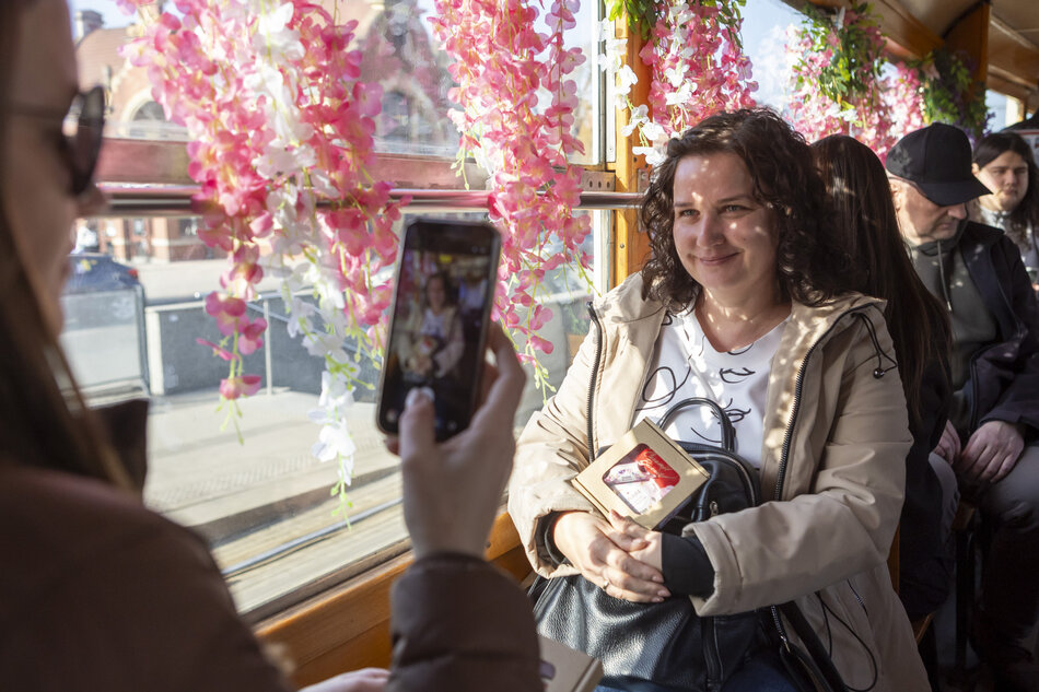 Na zdjęciu widać uśmiechniętą kobietę siedzącą w udekorowanym kwiatami pojeździe, której inna osoba robi zdjęcie telefonem komórkowym. W tle, za oknem i wewnątrz kabiny, dostrzec można obfite girlandy różowych kwiatów oraz innych pasażerów.