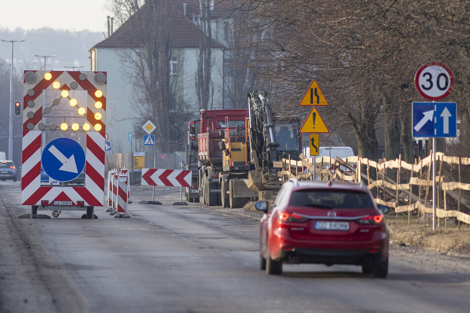 na zdjęciu samochód osobowy czerwony, jedzie ulicą, po lewej duży znak drogowy biało czerwony, po prawej drewniane ogrodzenie przy ulicy