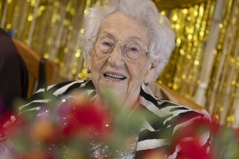 na pierwszym planie uśmiechnięta seniorka w okularach trzymająca kwiaty, które na zdjęciu są rozmazane. Z tyłu widnieje błyszcząca, złota oprawa