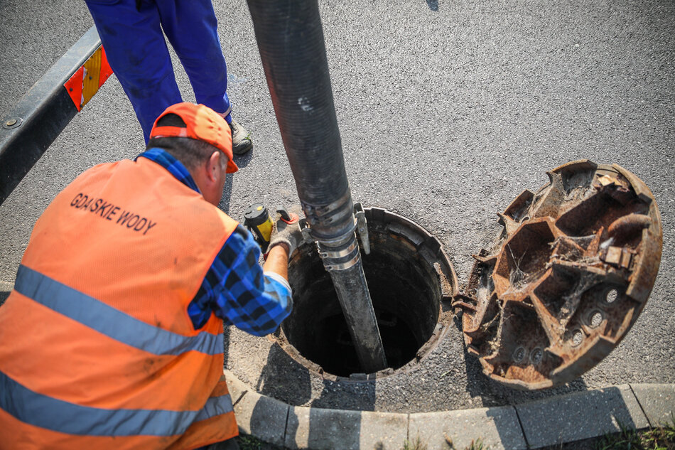 Pracownik spółki Gdańskie Wody w pomarańczowej kamizelce odblaskowej nadzoruje pracę rury ssącej wprowadzonej do otwartej studzienki kanalizacyjnej. Na drugim planie widać uniesioną, żeliwną pokrywę włazu oraz nogi drugiego pracownika w niebieskim stroju roboczym. 
