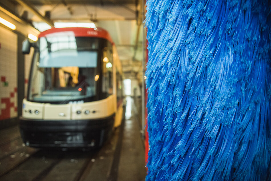 Na pierwszym planie widać zbliżenie na pionową, niebieską szczotkę obrotową, która jest częścią automatycznej myjni dla pojazdów wielkogabarytowych. W tle, w głębi hali, znajduje się nowoczesny, niskopodłogowy tramwaj oczekujący na czyszczenie.