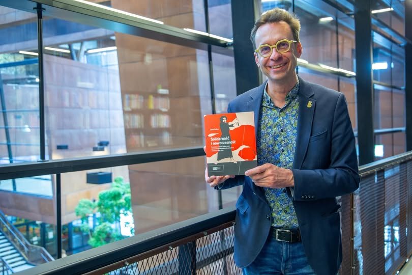 Szczupły mężczyzna w okularach pozuje z książką do zdjęcia 