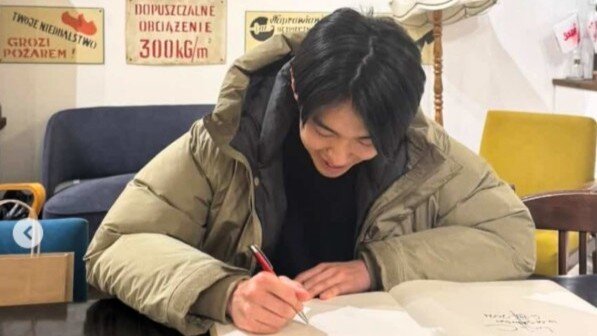 mężczyzna z długimi włosami siedzi przy stole i z uśmiechem na twarzy pisze coś w księdze. Ma na sobie kurtkę, z tyłu jest szara sofa. Wygląda jakby siedział w ciasnym pomieszczeniu