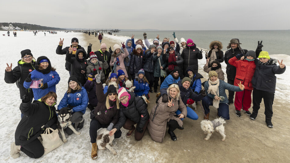 Spacer socjalizacyjny na plaży w Brzeźnie. Grupowe zdjęcie uczestników wraz z psami ze schroniska Promyk. Grupa uśmiechniętych ludzi macha do osoby wykonującej fotografię.