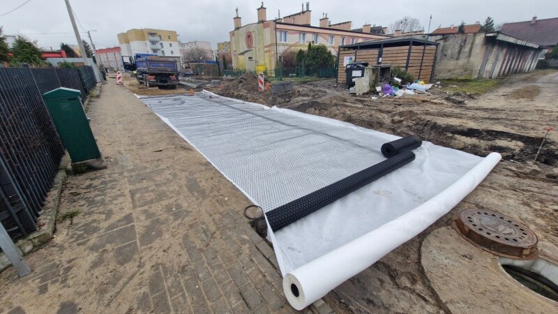 na zdjęciu rodzaj jasnej folii, bardzo szerokiej ułożonej na jezdni, widać dużo piasku, rozkopaną ulicę i koparkę