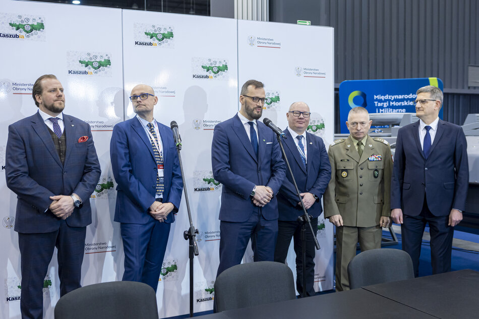 Grupa sześciu mężczyzn stojących przed tłem z logo i tekstem, wszyscy ubrani w formalny strój biznesowy i wydają się być na konferencji prasowej lub wydarzeniu, mężczyzna w środku mówi do mikrofonu.