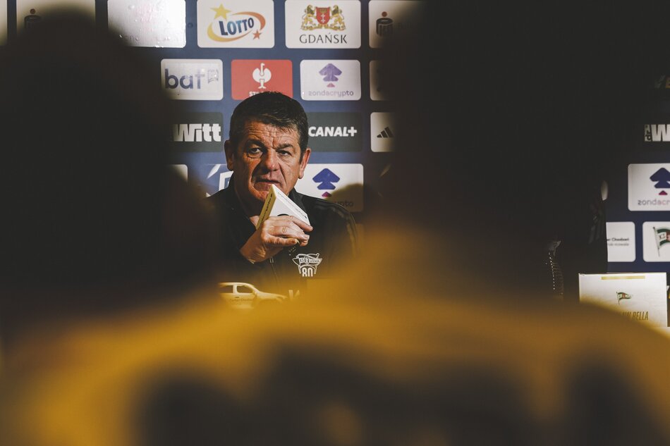 Zdjęcie mężczyzny w wieku lat około 60, siedzi w dresie, przy stole, w ręce trzyma mikrofon 