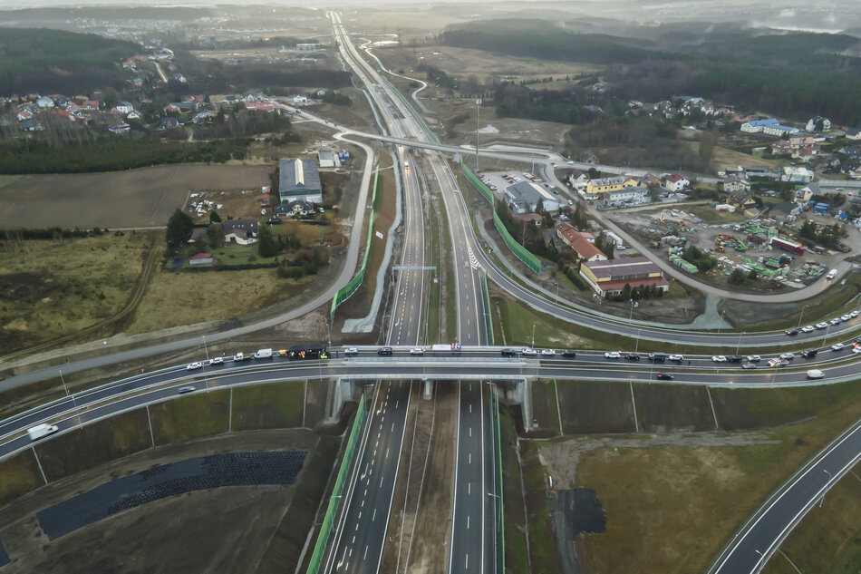 Na zdjęciu widać z lotu ptaka rozbudowany węzeł drogowy z nowoczesną drogą ekspresową oraz przebiegającym nad nią wiaduktem. Wokół trasy znajdują się tereny zabudowane, pola oraz place budowy, co wskazuje na niedawno zakończoną lub trwającą inwestycję infrastrukturalną. 