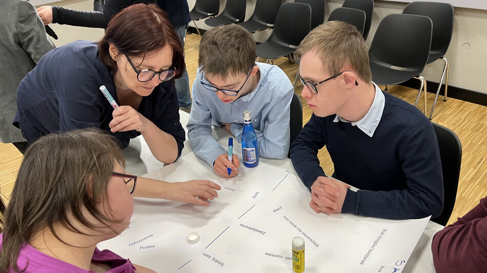 trzy osoby w młodym wieku siedzą przy stole, na nim stos kartek. Obok nich stoi kobieta i wskazuje na kartki, wygląda jakby coś tłumaczyła 