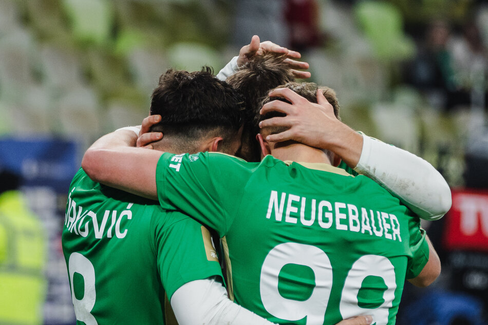 Trzej zawodnicy w biało-zielonych strojach obejmują się przyjaźnie po zdobyciu bramki 