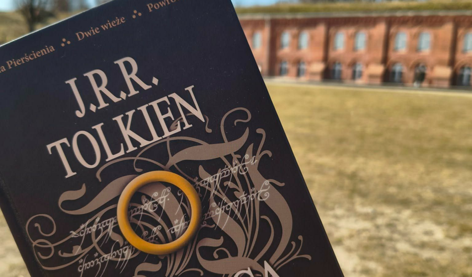 Zdjęcie przedstawia polskie wydanie książki J.R.R. Tolkiena, na której okładce położono żółty pierścień nawiązujący do słynnego Jedynego Pierścienia. W tle widoczny jest zabytkowy, ceglany budynek, co nadaje całości klimatyczny, historyczny charakter. 